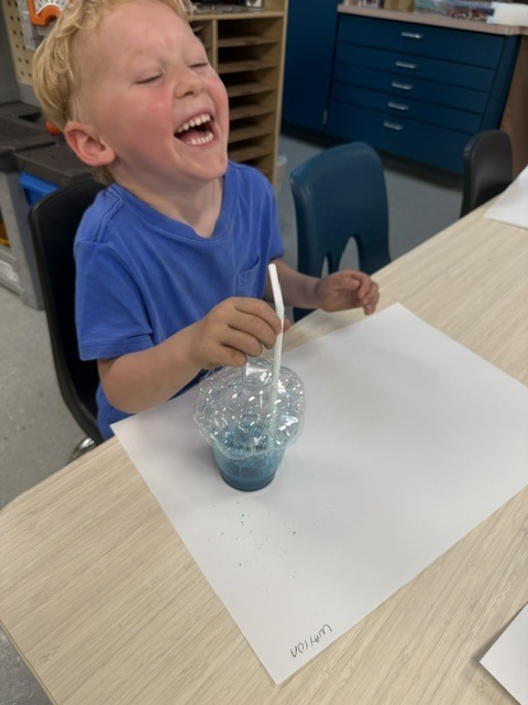 A laughing preschooler holds a straw beside a cup covered in bubbles after completing a colorful bubble science experiment.