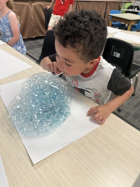 A smiling preschooler blows through a straw to create a large mound of blue bubbles during a classroom science activity.