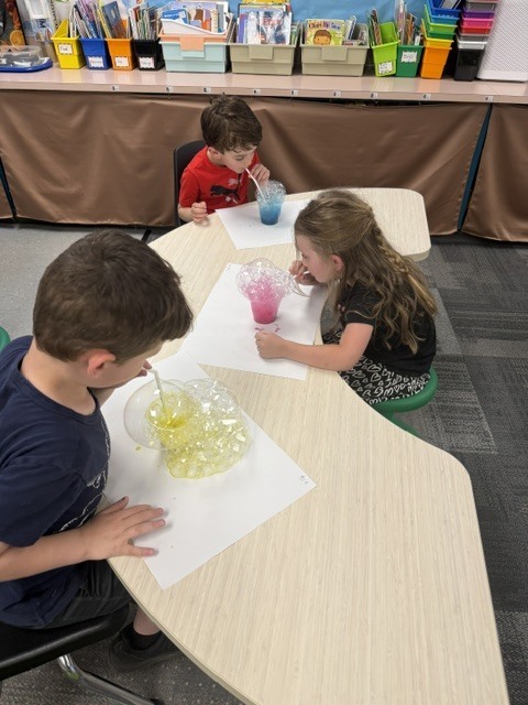 Three preschoolers blow through straws into cups of colorful bubble mixtures, watching bubbles overflow onto paper during a classroom science exploration.