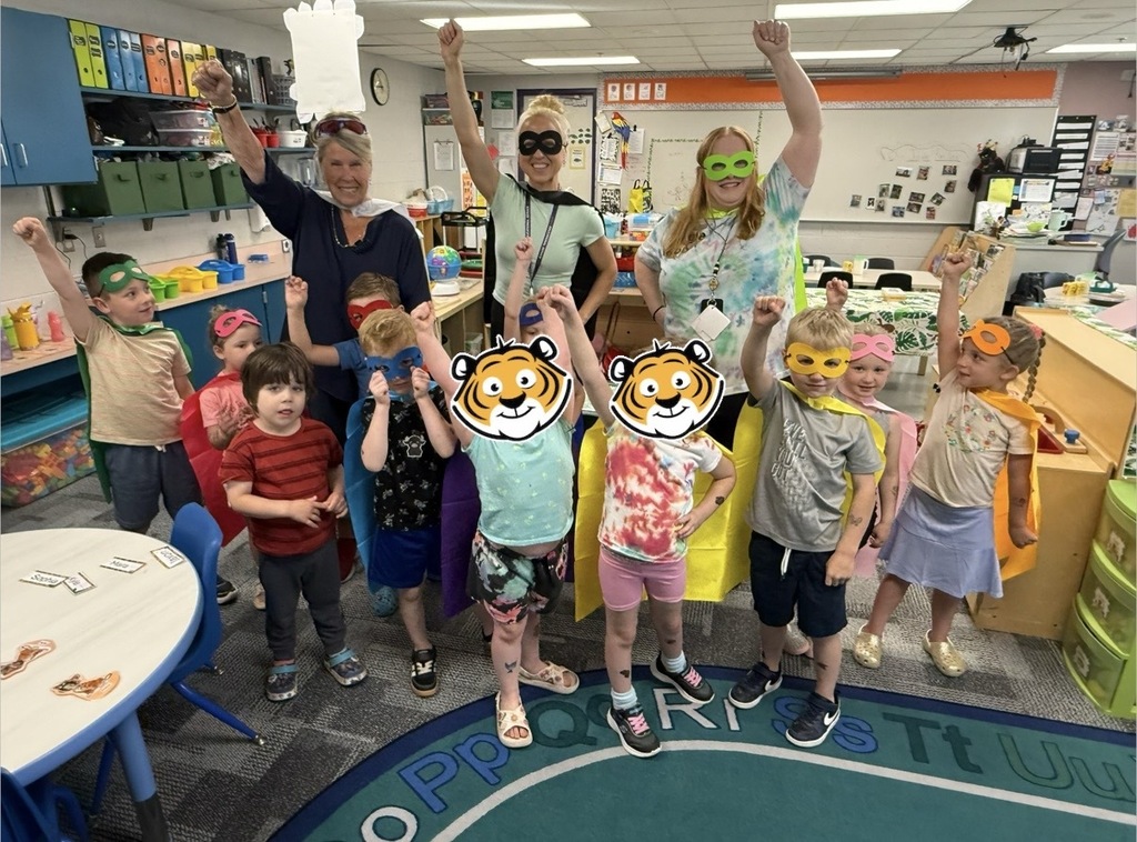 Preschool children and teachers in the Jungle Room classroom dressed as superheroes for Superhero Day. The children wear colorful masks and capes while posing with their arms raised in superhero stances. Three teachers stand behind them smiling and joining in the fun.