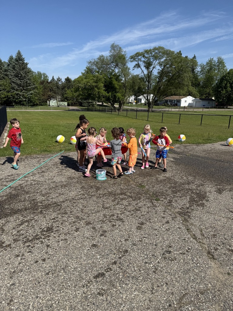 Preschool children playing outside during Water Day activities on a sunny day. Children wear swimsuits and summer clothes while exploring water tables, spray toys, beach balls, and buckets on the playground pavement.