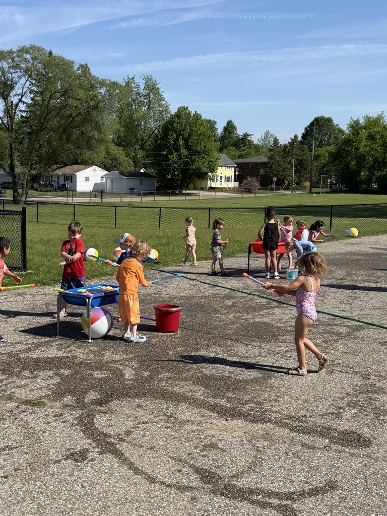 Preschool children playing outside during Water Day activities on a sunny day. Children wear swimsuits and summer clothes while exploring water tables, spray toys, beach balls, and buckets on the playground pavement.