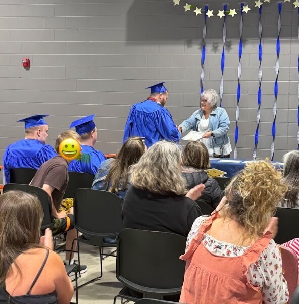 A student dressed in a blue graduation gown is shaking hands with a woman wearing a white top with a denim jacket