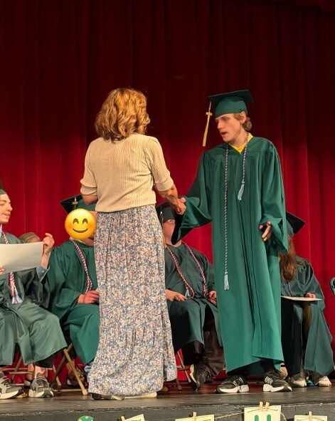 A student in a green graduation gown is shaking hands with a female adult in a floral skirt and tan shirt.