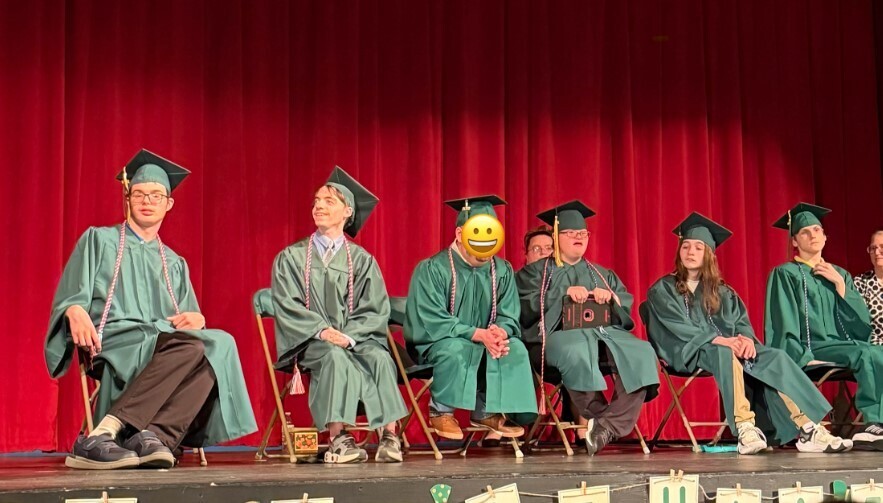 6 students sit in chairs on a stage in green graduation gowns