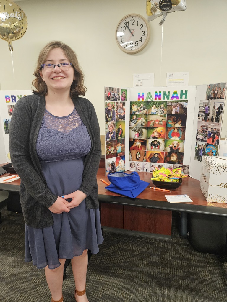 Hannah, wearing glasses, a blue dress, and a grey cardigan, smiles in front of her graduation tri-fold board covered in childhood and family photos.