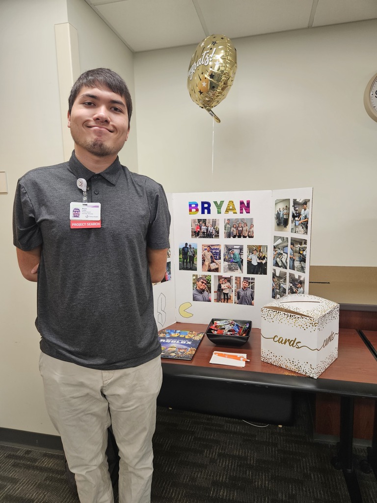 Bryan stands proudly with his hands behind his back, wearing a grey polo with a Project SEARCH ID badge next to his graduation display board.