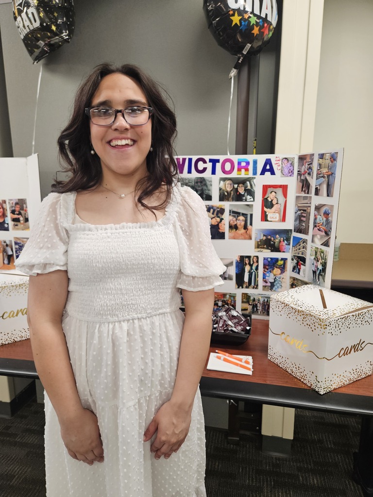 Victoria, wearing glasses and a white textured dress, smiles in front of her graduation board decorated with photos of friends and activities.