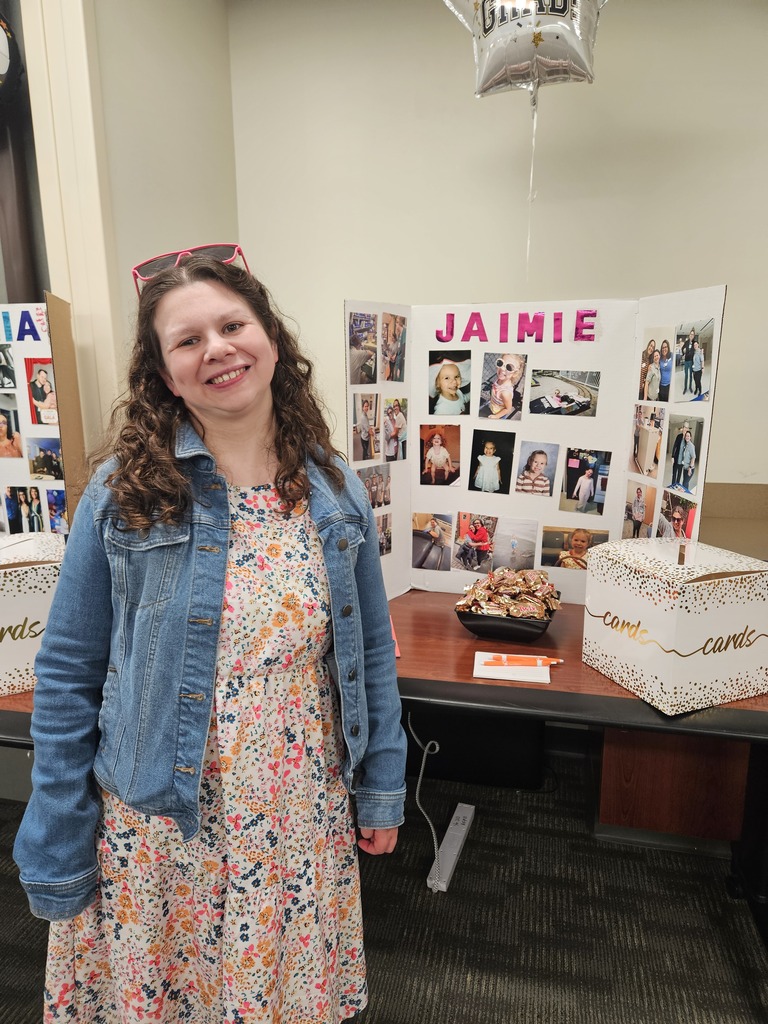 Jaimie smiles warmly while wearing a floral dress and a denim jacket, standing beside her graduation display filled with personal photos