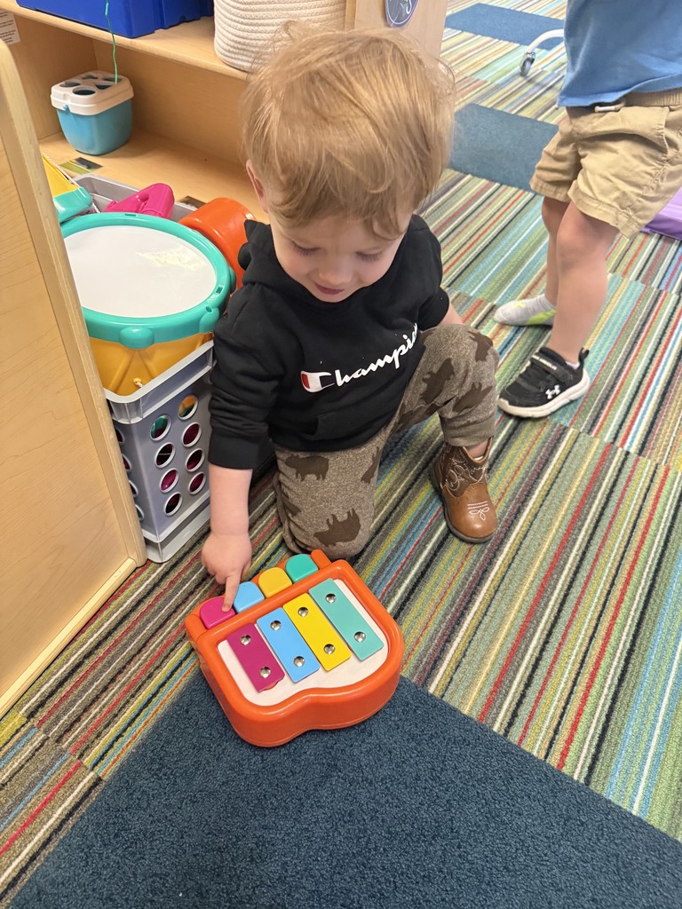 An Early Head Start child kneels on a classroom carpet while pressing a colorful toy xylophone. Musical instruments and classroom materials are nearby as the child explores sound and play.