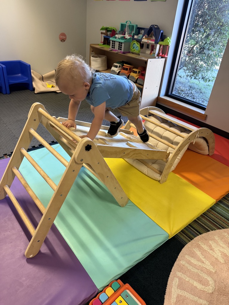 An Early Head Start child climbs across a wooden indoor climbing structure placed on colorful mats. The child is focused on balancing and exploring movement in a classroom gross motor area.
