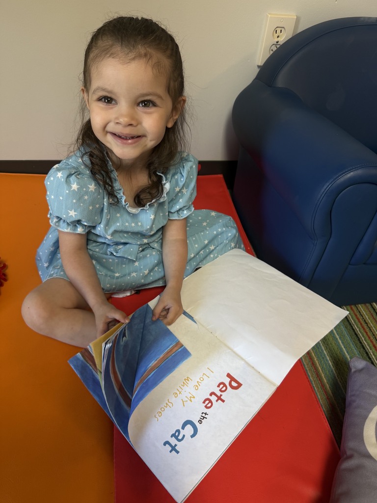 An Early Head Start child sits on a colorful mat in a classroom reading area, smiling while looking through a picture book. A child-sized chair and soft seating are nearby, creating a cozy space for exploring books and early literacy.