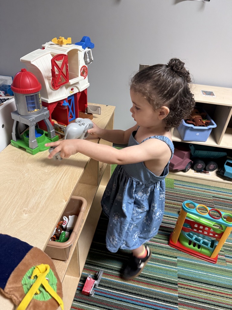 An Early Head Start child stands at a play shelf, exploring a toy barn and animal figures. The child is engaged in imaginative play while interacting with classroom toys in a learning center.