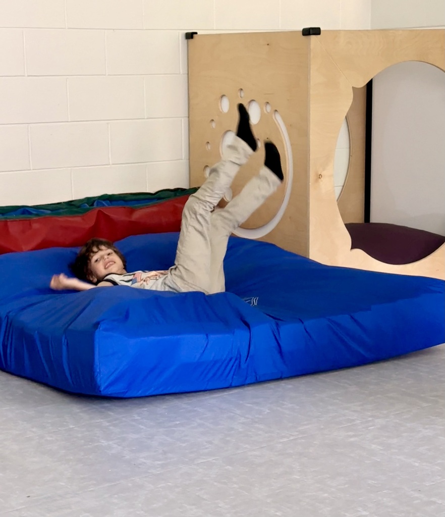 A smiling young boy happily kicks his legs up in the air while lying on a large, blue crash mat in a sensory  room.