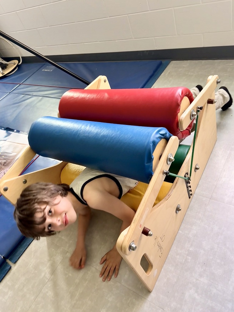A young boy smiles at the camera as he crawls through a wooden sensory steamroller squeeze machine with large red and blue padded rollers.
