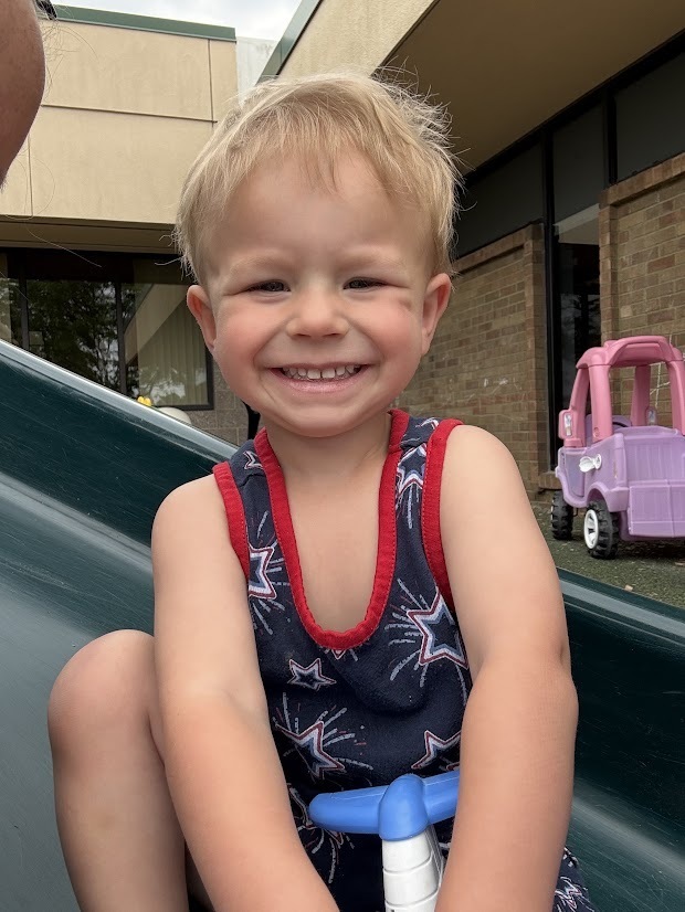 A smiling young child sits on a playground slide wearing a patriotic romper, with outdoor play equipment in the background.