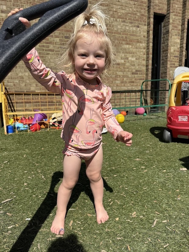 A young child in a pink flamingo swimsuit smiles while standing barefoot outdoors and holding onto playground equipment.