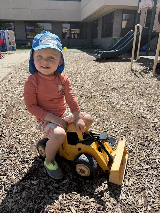 A young child wearing a blue sun hat sits on a yellow toy construction vehicle on a woodchip playground, smiling at the camera.
