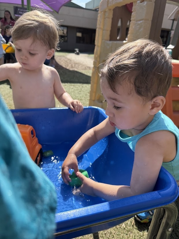 Two young children play at a blue water table outdoors, splashing water and exploring floating toys during Water Day.