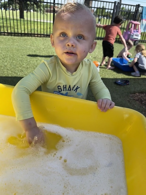 A young child in a yellow swim shirt smiles while exploring a tub of soapy water during Water Day, with classmates playing nearby.