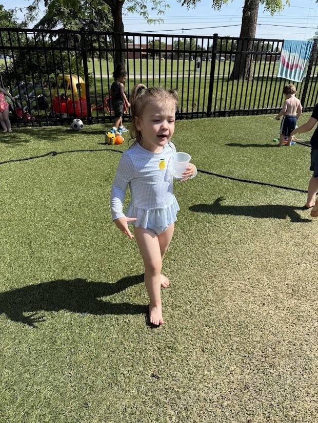 A young child wearing a light blue swimsuit walks across the playground holding a cup during an outdoor Water Day activity.