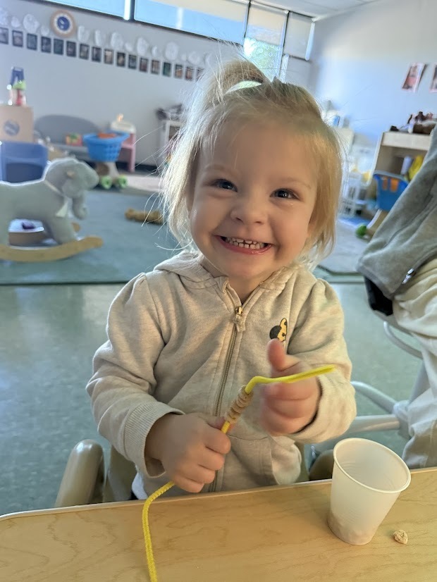 A smiling young child proudly holds a partially completed bird feeder made from cereal threaded onto a pipe cleaner.