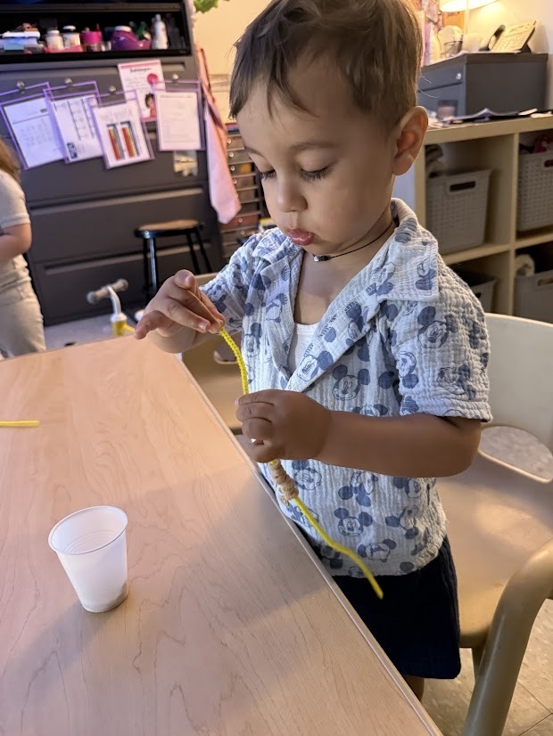 A young child carefully threads cereal onto a pipe cleaner while making a bird feeder at a classroom table.