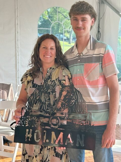A smiling woman and a young man posing together inside a tenet while holding a custom metal cutout sign that features horses and the name "Holman."