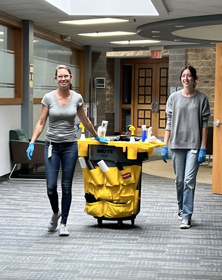 Two women with a bin on wheels walking down the hall. 