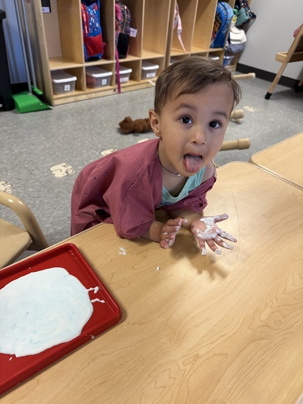 A child smiles at the camera with their tongue out while exploring white oobleck on a table, hands covered in the sensory mixture.