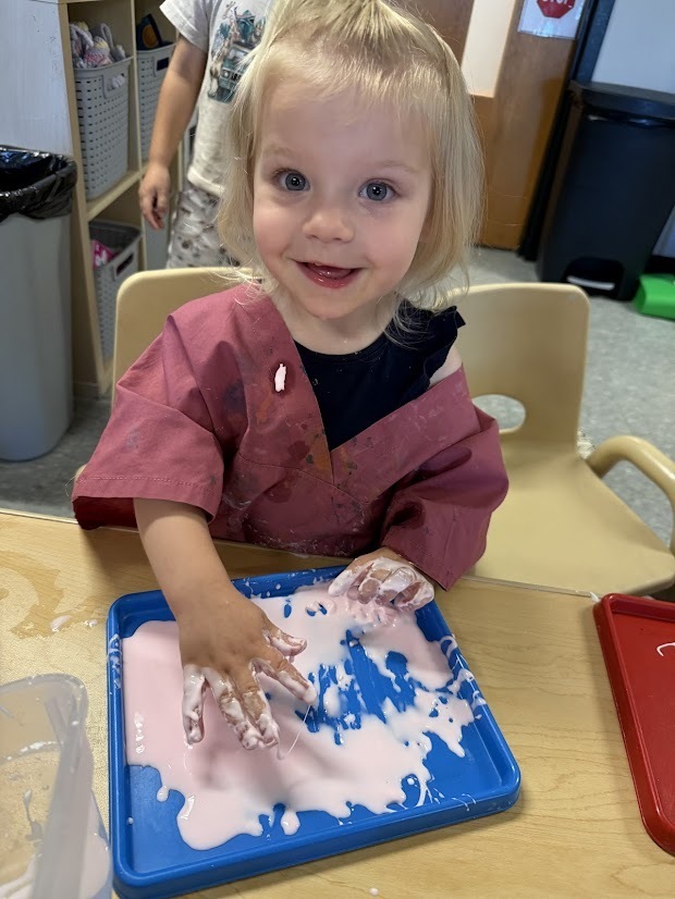 A smiling child presses both hands into a tray of pink oobleck, enjoying the messy sensory experience during classroom exploration.