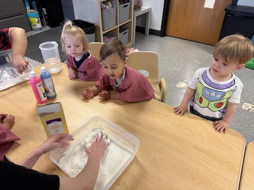 Several toddlers gather around a table, watching a bin of oobleck ingredients as they explore the changing texture together.