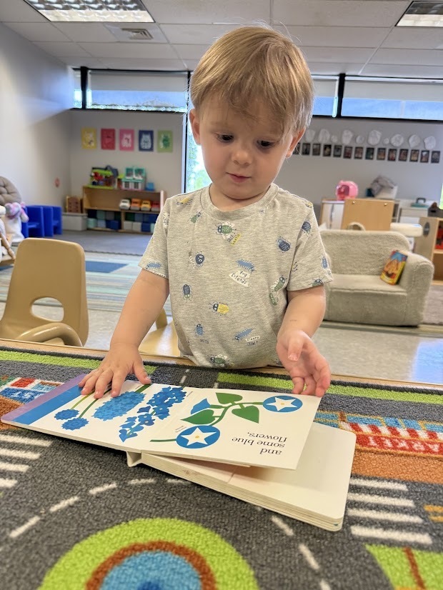 A young child stands at a classroom table, turning the pages of a picture book while exploring colorful illustrations.
