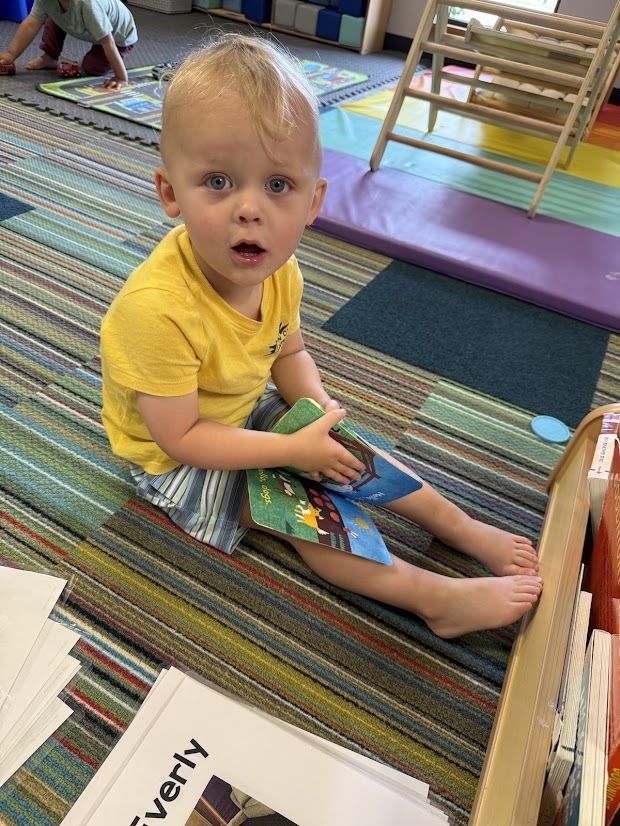 A child sits on a classroom rug holding a board book and looking toward the camera.