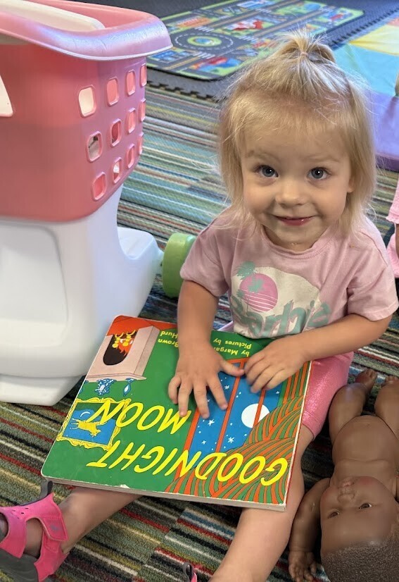A smiling child sits on a classroom rug holding a book.