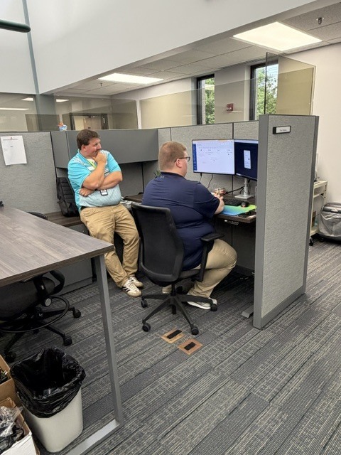 An indoor office setting showing a cubicle workspace. A man wearing a dark blue shirt and glasses sits in an office chair, facing two computer monitors displaying data and software interfaces. Another man, wearing a bright turquoise polo shirt and khaki pants, leans back against an adjacent desk with his arms crossed, thoughtfully looking over at the monitors. The room features grey carpet tiles, cubicle dividers with glass privacy panels, and standard office lighting.