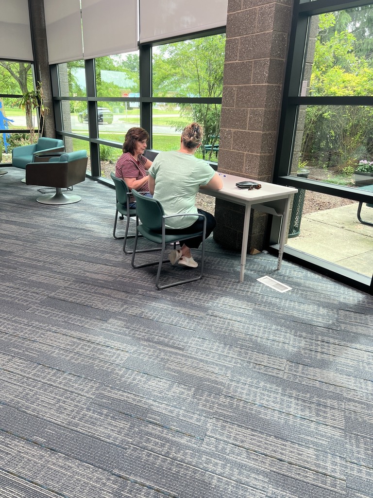 An indoor view of a brightly lit room featuring large floor-to-ceiling windows that overlook trees and a walkway outside. Two women are seated in teal chairs at a small rectangular table placed against a brick pillar. One woman wears a red-and-white striped shirt, and the other, viewed from behind, wears a light green t-shirt with her hair up in a claw clip. The room has textured blue and grey carpeting, with additional teal armchairs visible in the background.