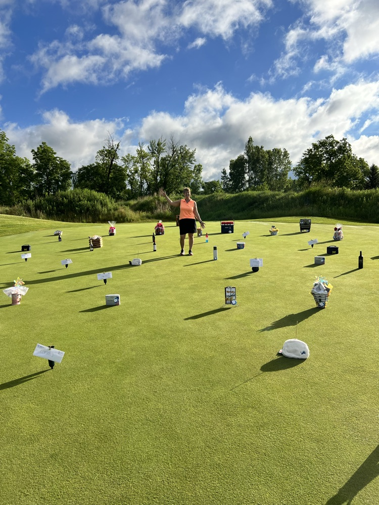 Adult female stands in middle of putting green surrounded with prizes. 