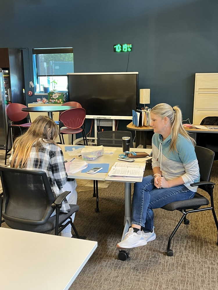 Two women sitting at a table one reading the other looks on.