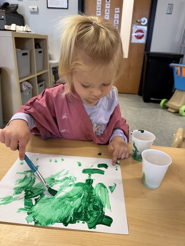 Young child painting with green paint on white paper using a paintbrush at a classroom table.