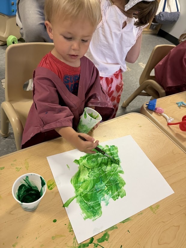 Young child painting with green paint on white paper using a paintbrush at a classroom table.