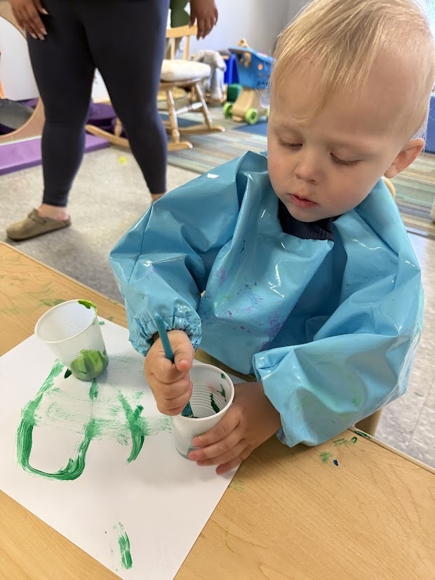 Young child painting with green paint on white paper using a paintbrush at a classroom table.