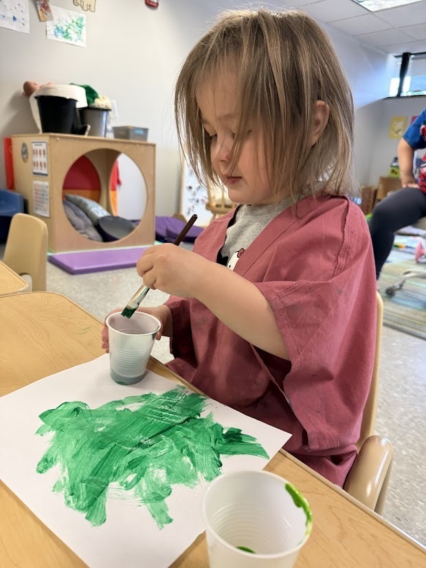 Young child painting with green paint on white paper using a paintbrush at a classroom table.