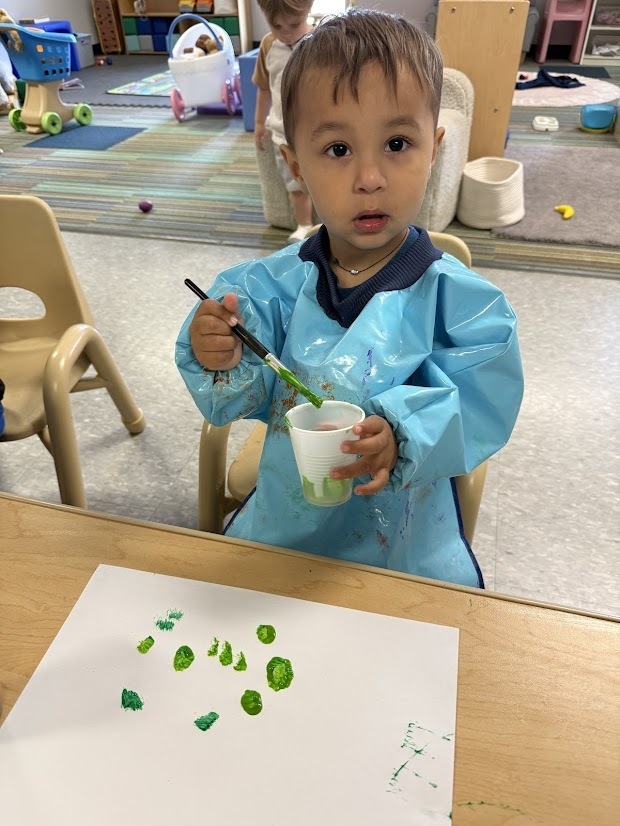 Young child painting with green paint on white paper using a paintbrush at a classroom table.