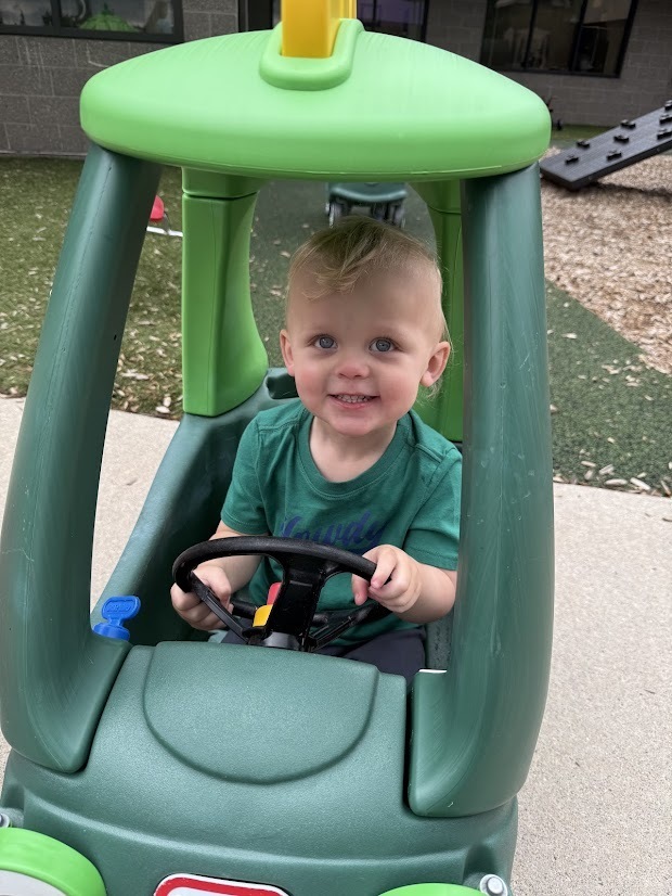 Young child smiling while sitting in a toy car and holding the steering wheel on the playground.
