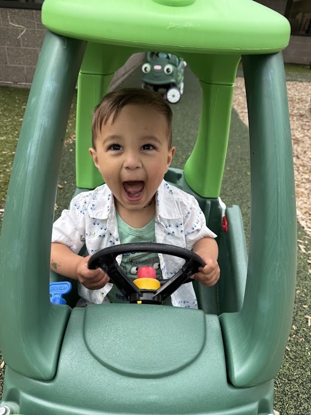 Young child smiling while sitting in a toy car and holding the steering wheel on the playground.