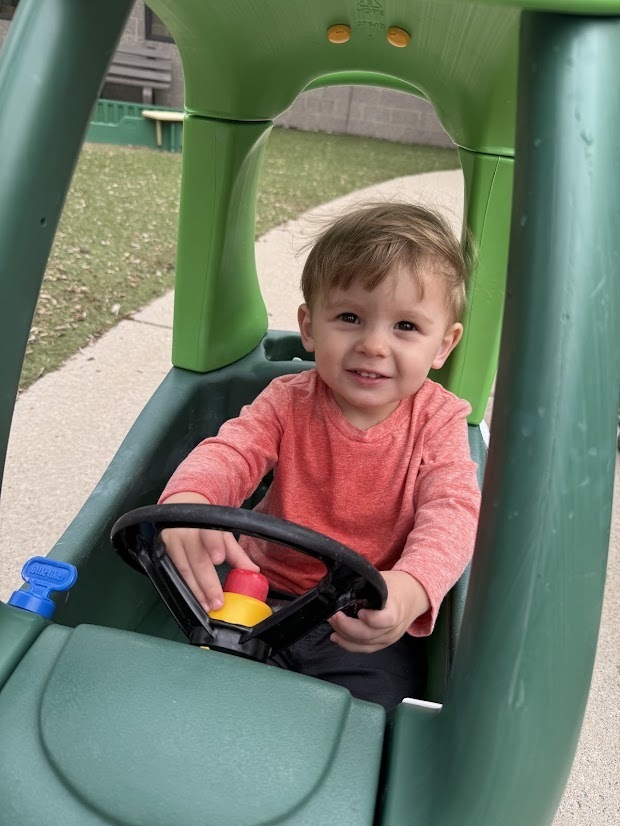 Young child smiling while sitting in a toy car and holding the steering wheel on the playground.