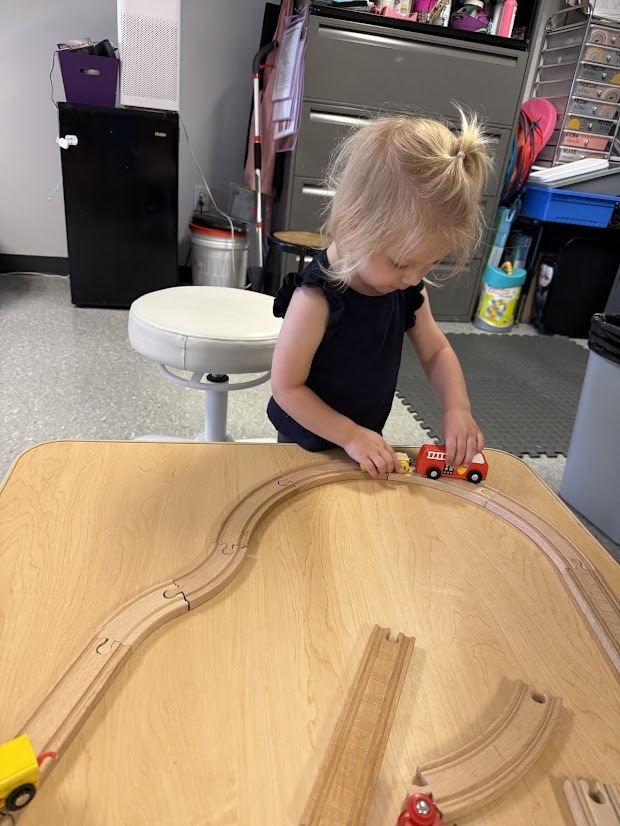 Young child arranges toy trains on a wooden track at a classroom table.