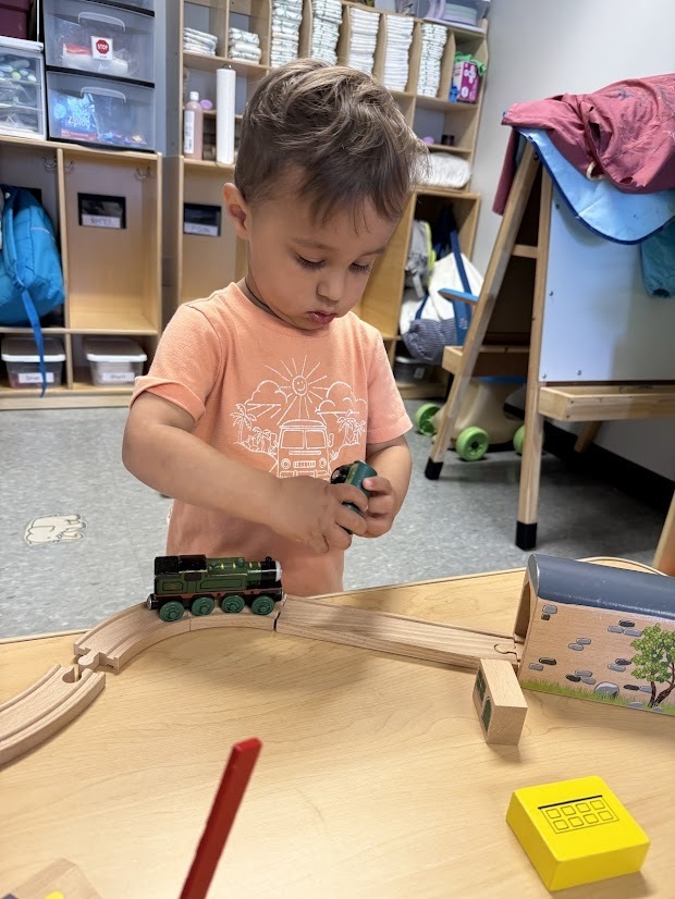 Young child focuses on connecting and moving toy trains along a wooden train track.