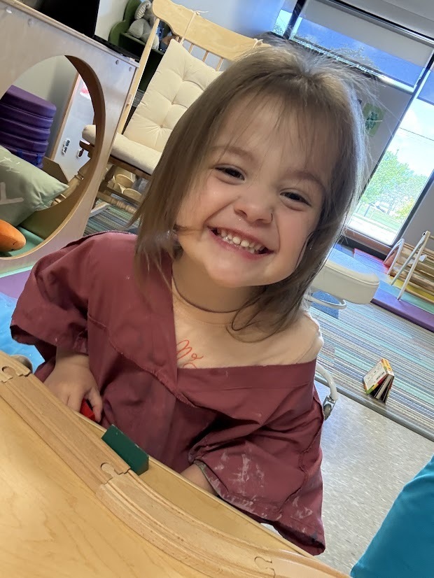 Young child smiling at the camera while sitting beside a wooden train track activity table.